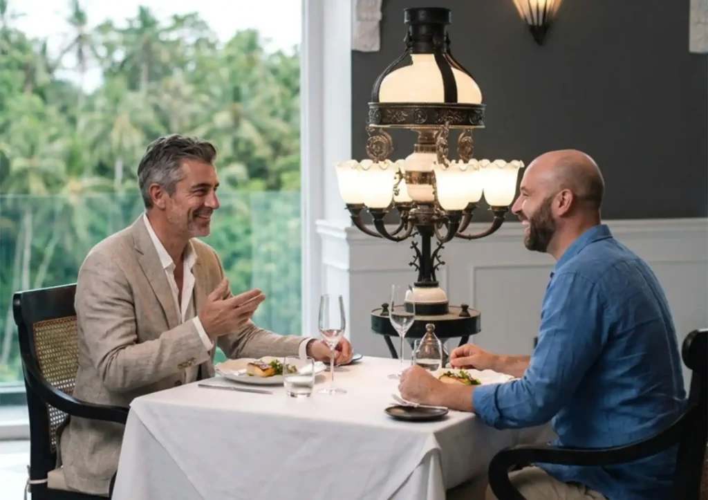 Two men laughing and conversing over an elegant meal beside an ornate table lamp, enjoying the atmosphere of fine dining Bali.