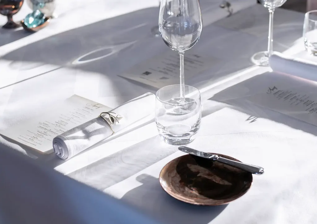 A sunlit table setting for fine dining ubud, featuring a printed menu, a rolled white napkin in a silver ring, clear glassware, and a rustic copper plate with a butter knife on a crisp white tablecloth.