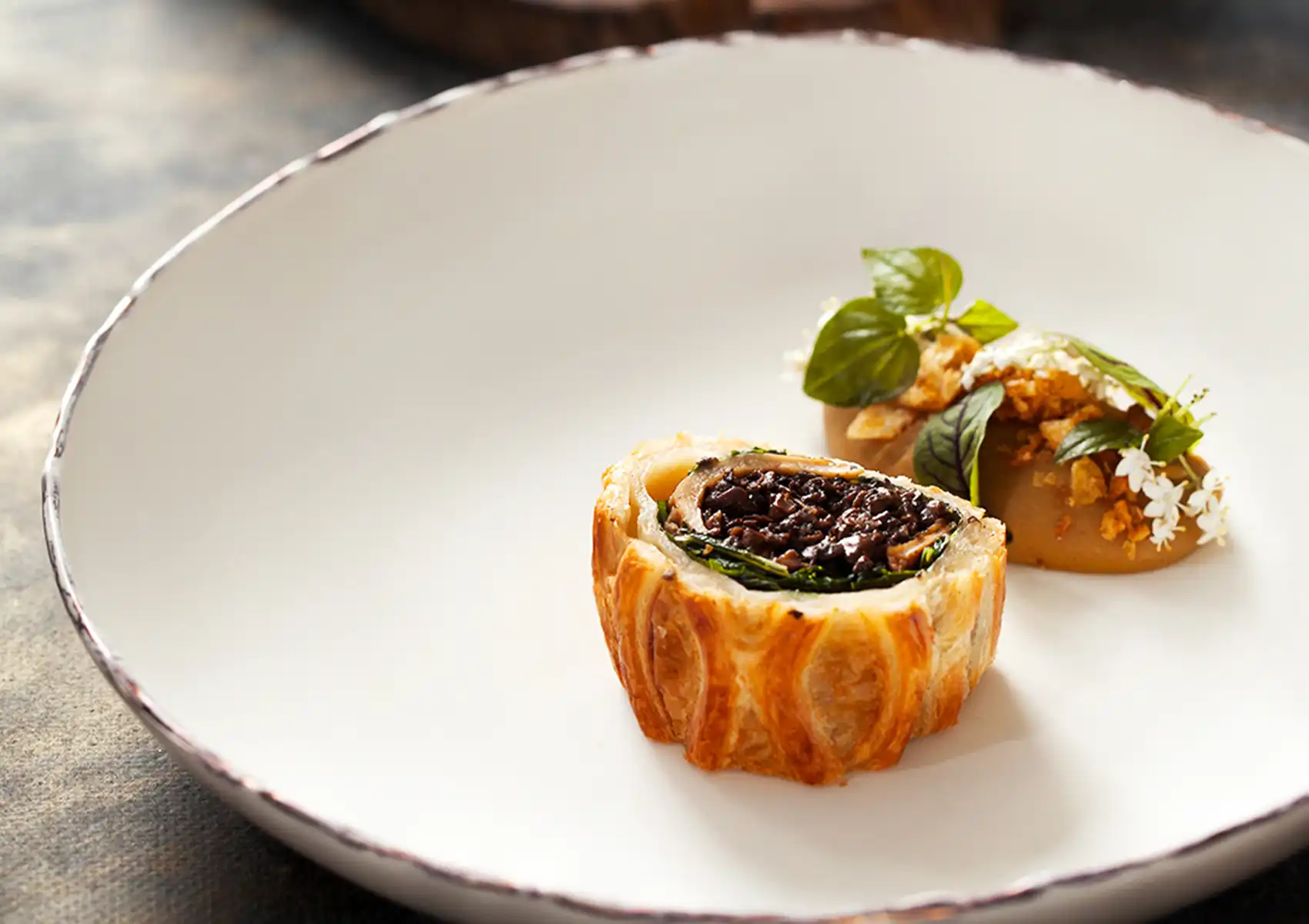 A beautifully plated slice of mushroom wellington with flaky pastry and dark mushroom filling, accompanied by an herb-garnished puree on a white plate, showcasing fine dining ubud.