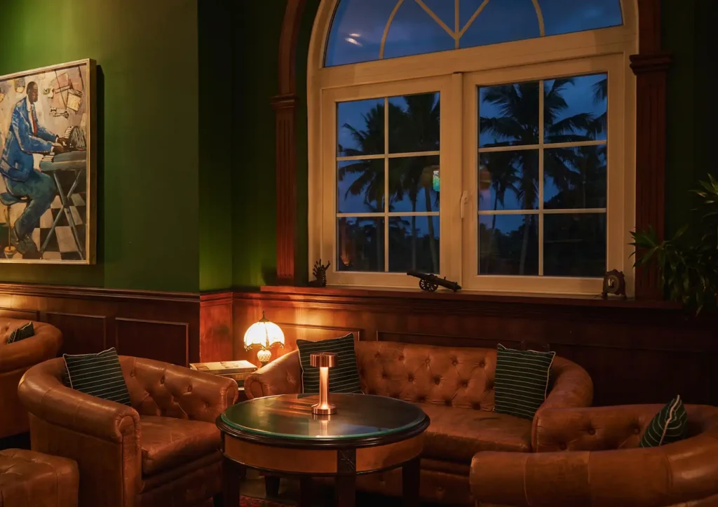 A sophisticated, dimly lit speakeasy lounge featuring dark green walls, wood paneling, tufted leather Chesterfield sofas, and a large arched window overlooking palm trees before an evening of fine dining Ubud