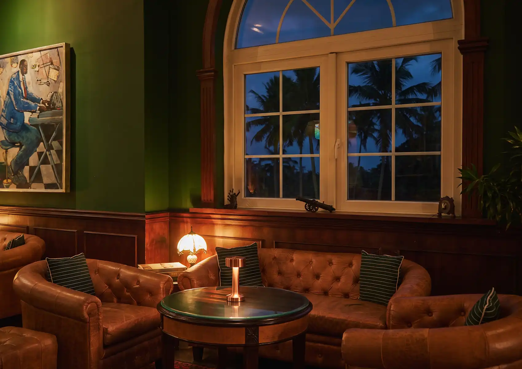 A sophisticated, dimly lit speakeasy lounge featuring dark green walls, wood paneling, tufted leather Chesterfield sofas, and a large arched window overlooking palm trees before an evening of fine dining Ubud
