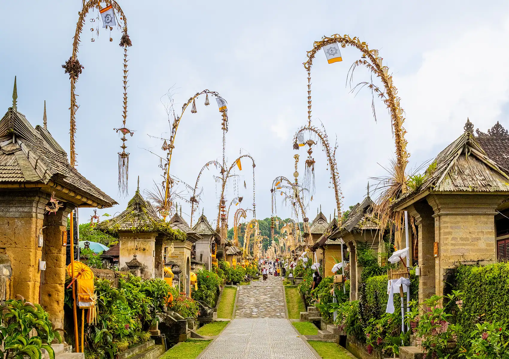 A scenic village stone pathway lined with traditional Balinese architecture and tall, decorative bamboo penjor poles, a beautiful area to explore before enjoying fine dining ubud.