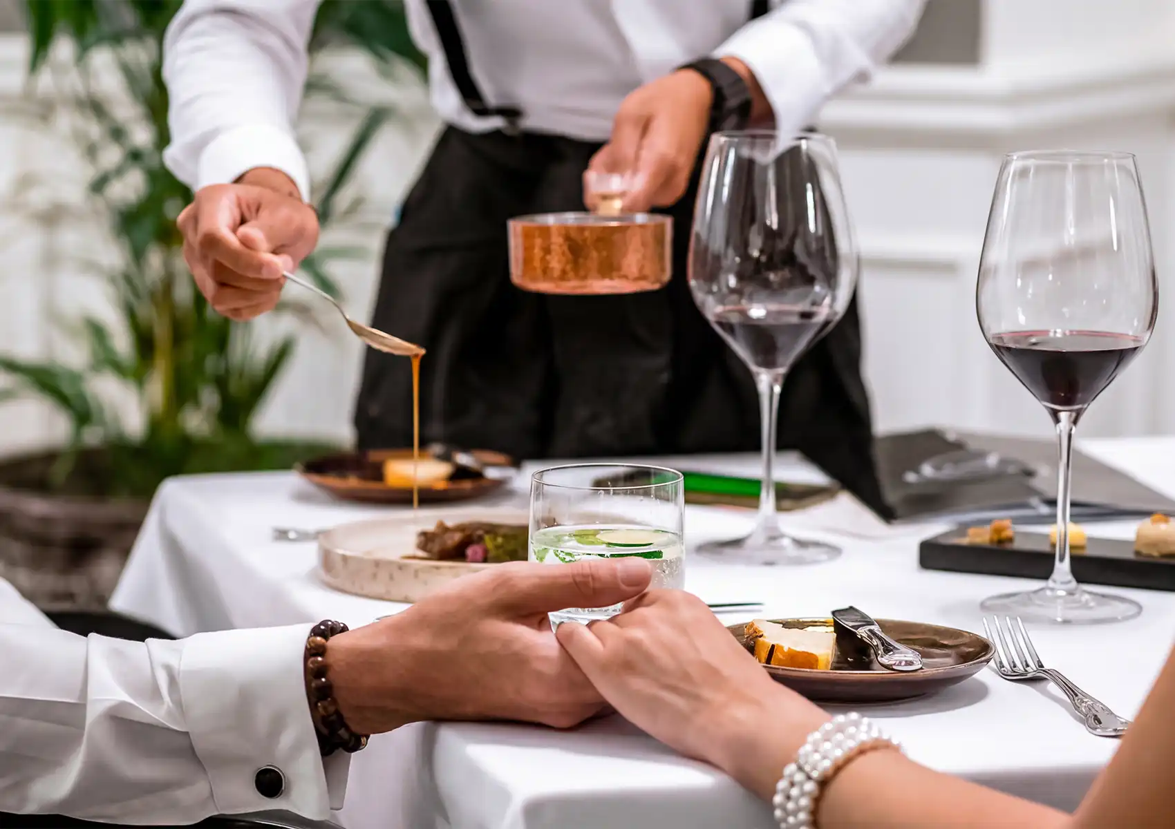 A close-up of a couple holding hands across a white-tablecloth table with glasses of red wine, while a waiter pours sauce from a copper pan during a meal at a fine dining Ubud restaurant.