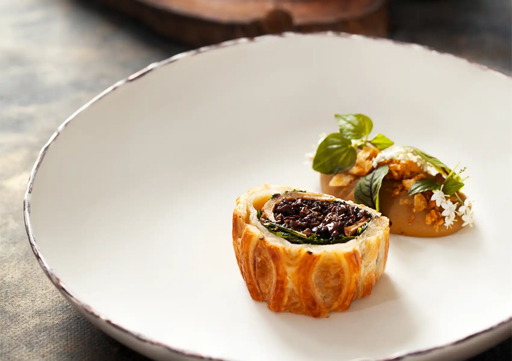 A beautifully plated slice of mushroom wellington with flaky pastry and a dark, rich filling, accompanied by an herb-garnished puree on a white plate, highlighting vegetarian fine dining ubud.