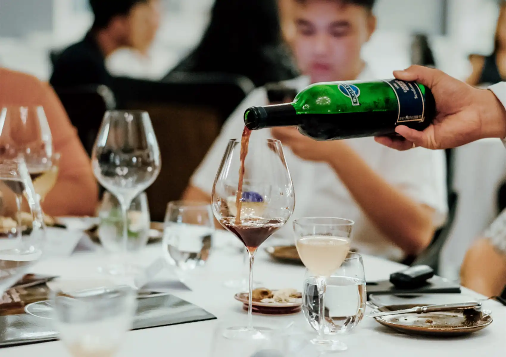 A close-up of a server pouring red wine from a green bottle into a large glass at a white-clothed table, capturing the experience of fine dining ubud.