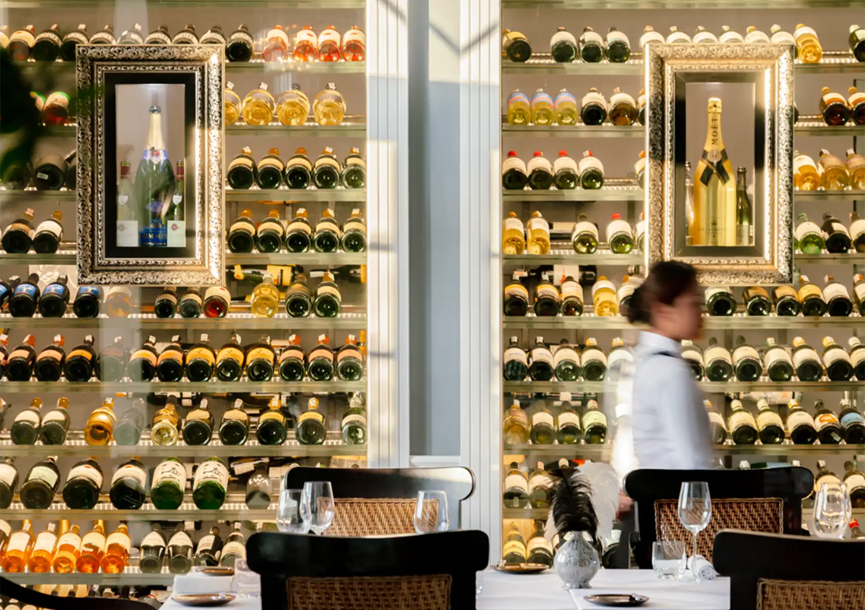 A server walks quickly past an expansive, warmly lit glass wine cellar featuring neatly organized bottles and ornate framed displays, setting the scene for fine dining bali.