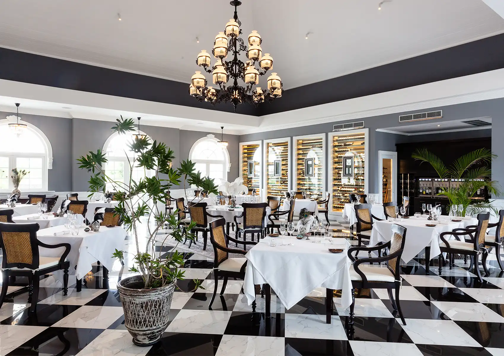 A spacious, elegant restaurant interior featuring a black and white checkered floor, arched windows, an ornate black chandelier, and beautifully set tables prepared for fine dining bali.
