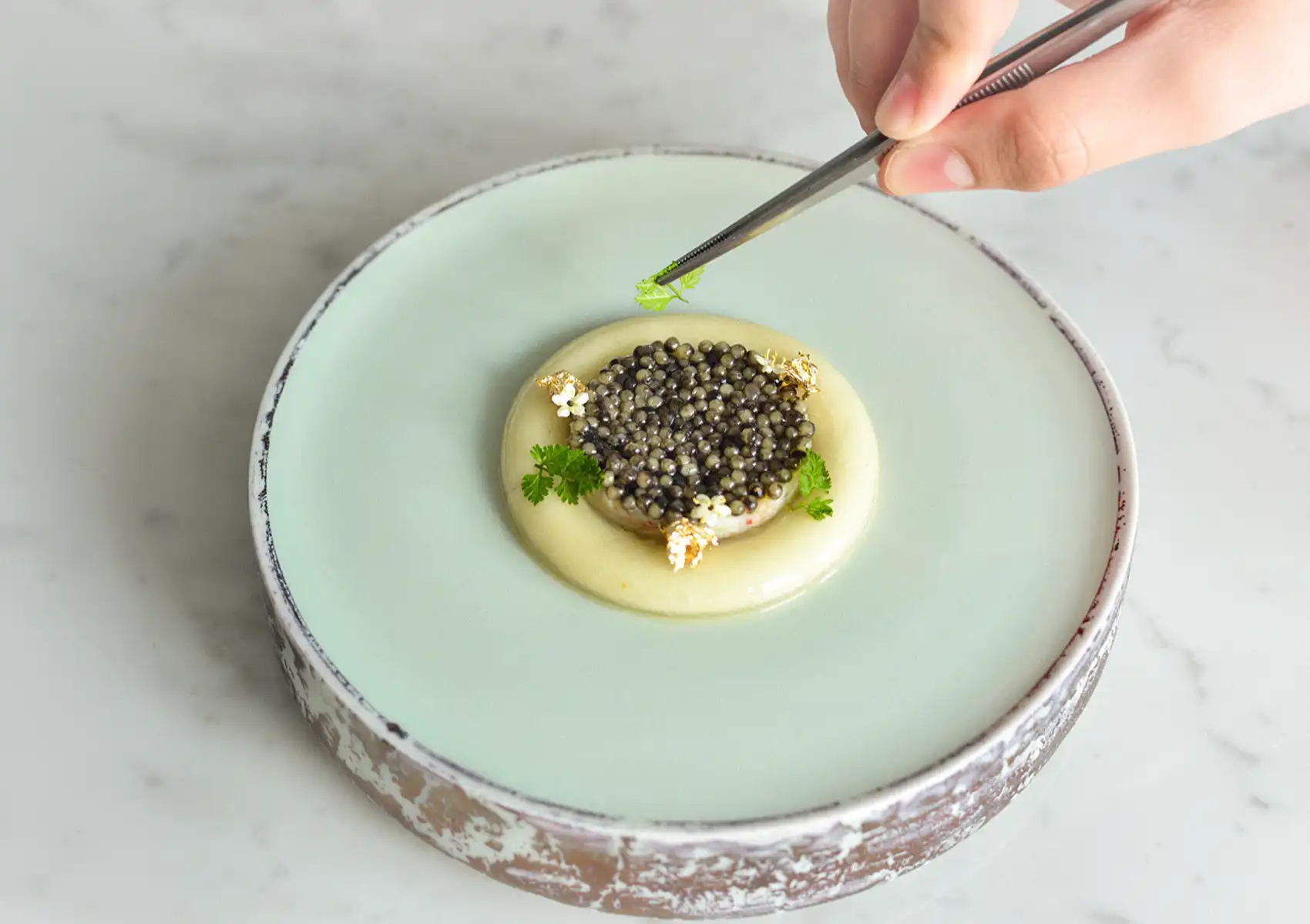 A chef's hand uses fine tweezers to precisely place a tiny green garnish onto an elegant caviar and cream dish, illustrating the meticulous culinary presentation expected in fine dining bali.