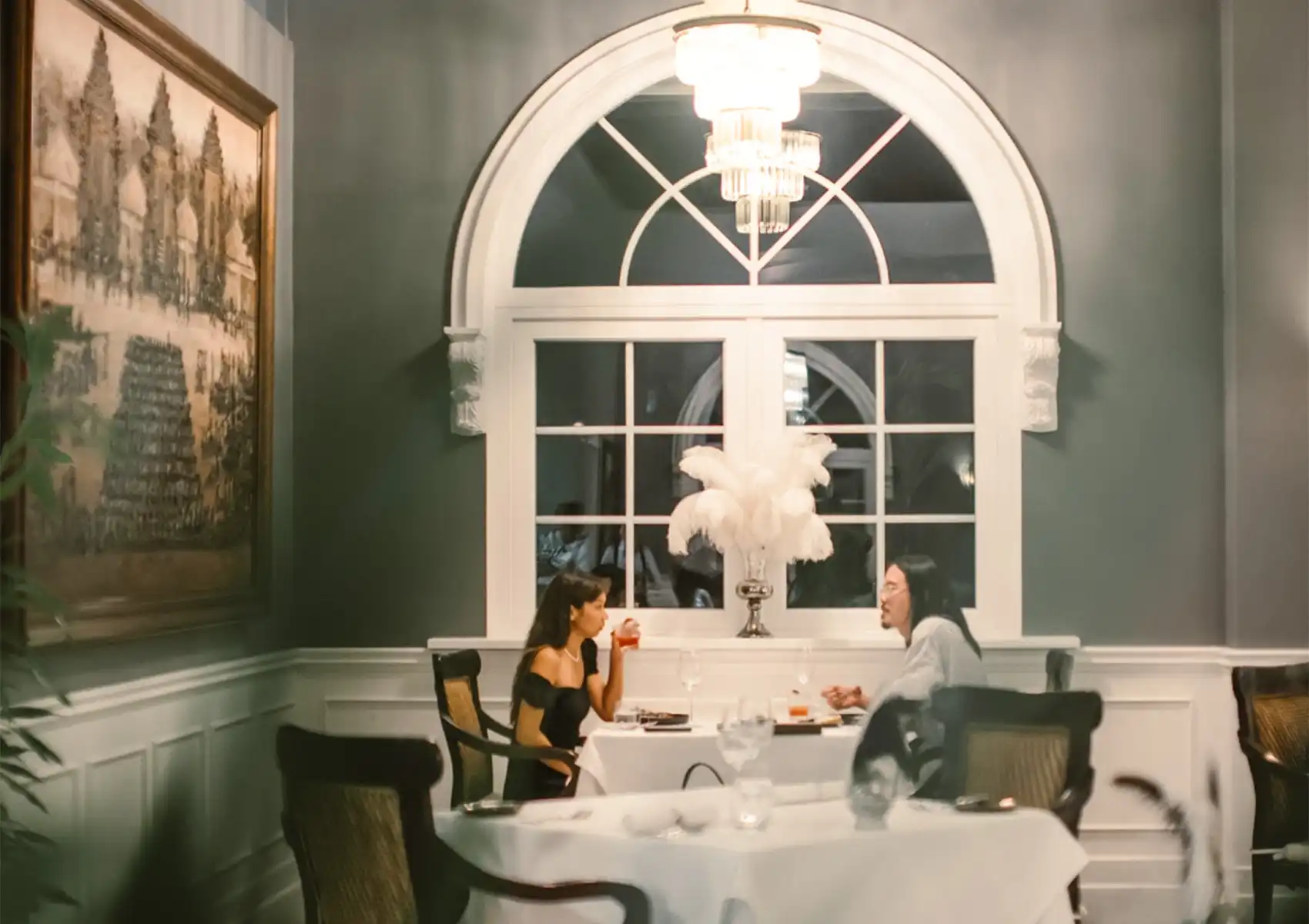 A couple sits at a beautifully set table near a large arched window, enjoying a conversation and drinks during a fine dining bali meal.