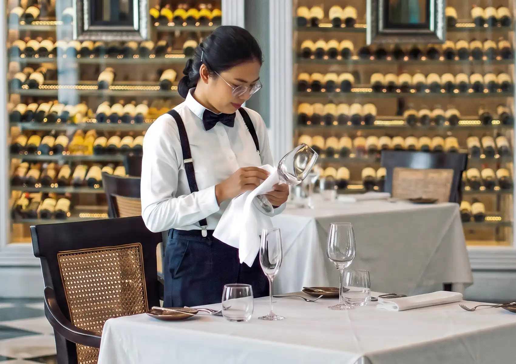 A server wearing a bowtie and suspenders carefully polishes a wine glass with a white cloth at a perfectly set table, showcasing the meticulous service of fine dining bali with a large wine cellar in the background.