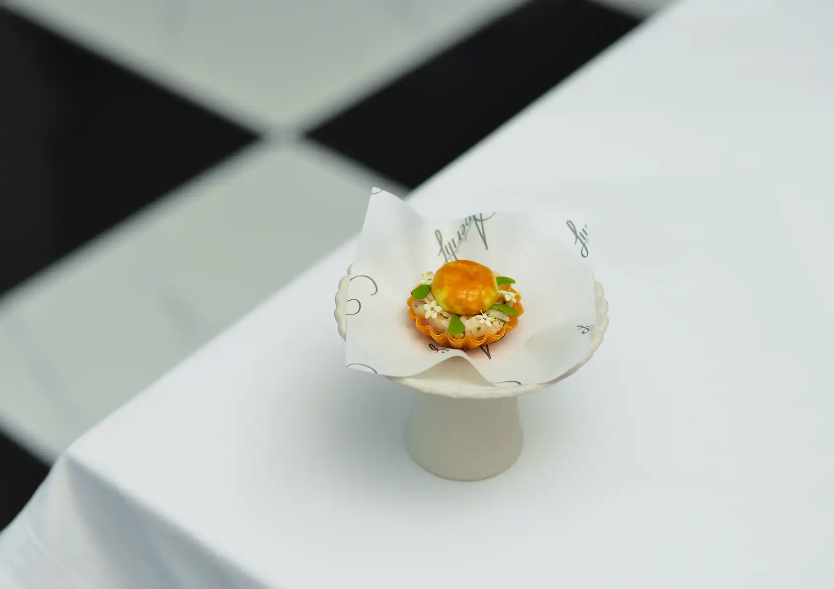 A delicate, bite-sized appetizer garnished with tiny white flowers sits on a small white pedestal over a white tablecloth, showcasing an intricate fine dining bali presentation.
