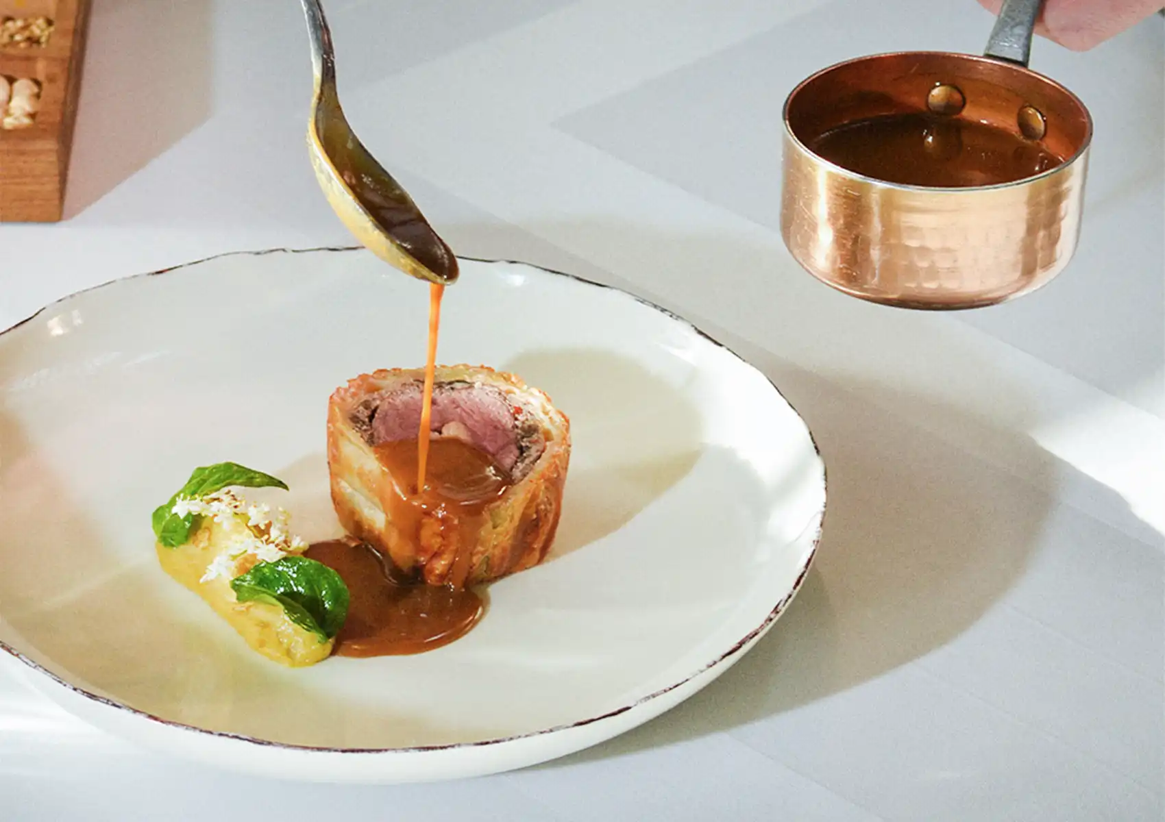Rich brown sauce is being poured from a copper pot over a plated slice of pastry-wrapped meat and a yellow garnish, showcasing a fine dining Bali experience.