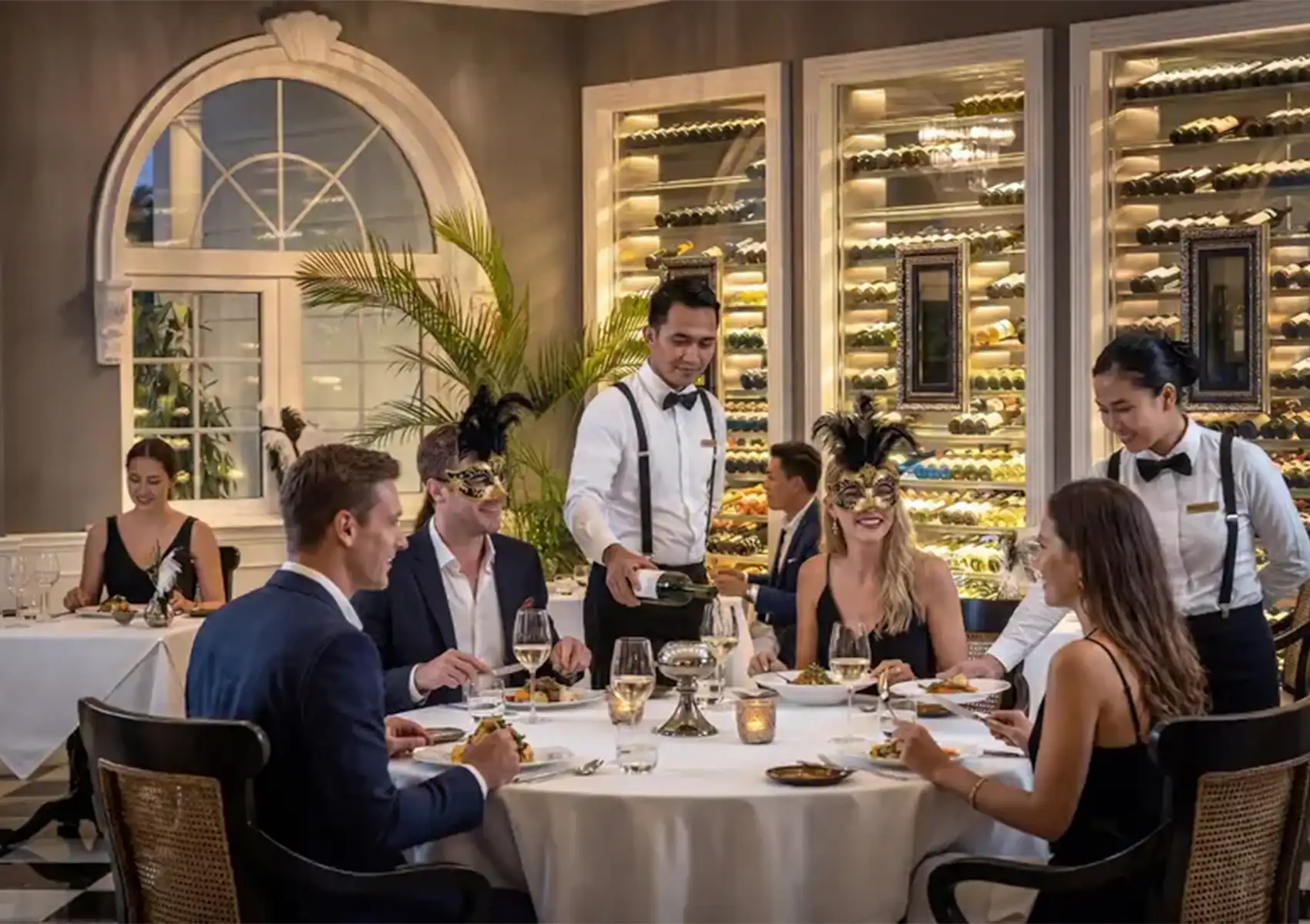 A waiter pours wine for formally dressed guests, some wearing feathered masks, seated at a round table in front of a glowing wine cellar during a festive fine dining bali experience.