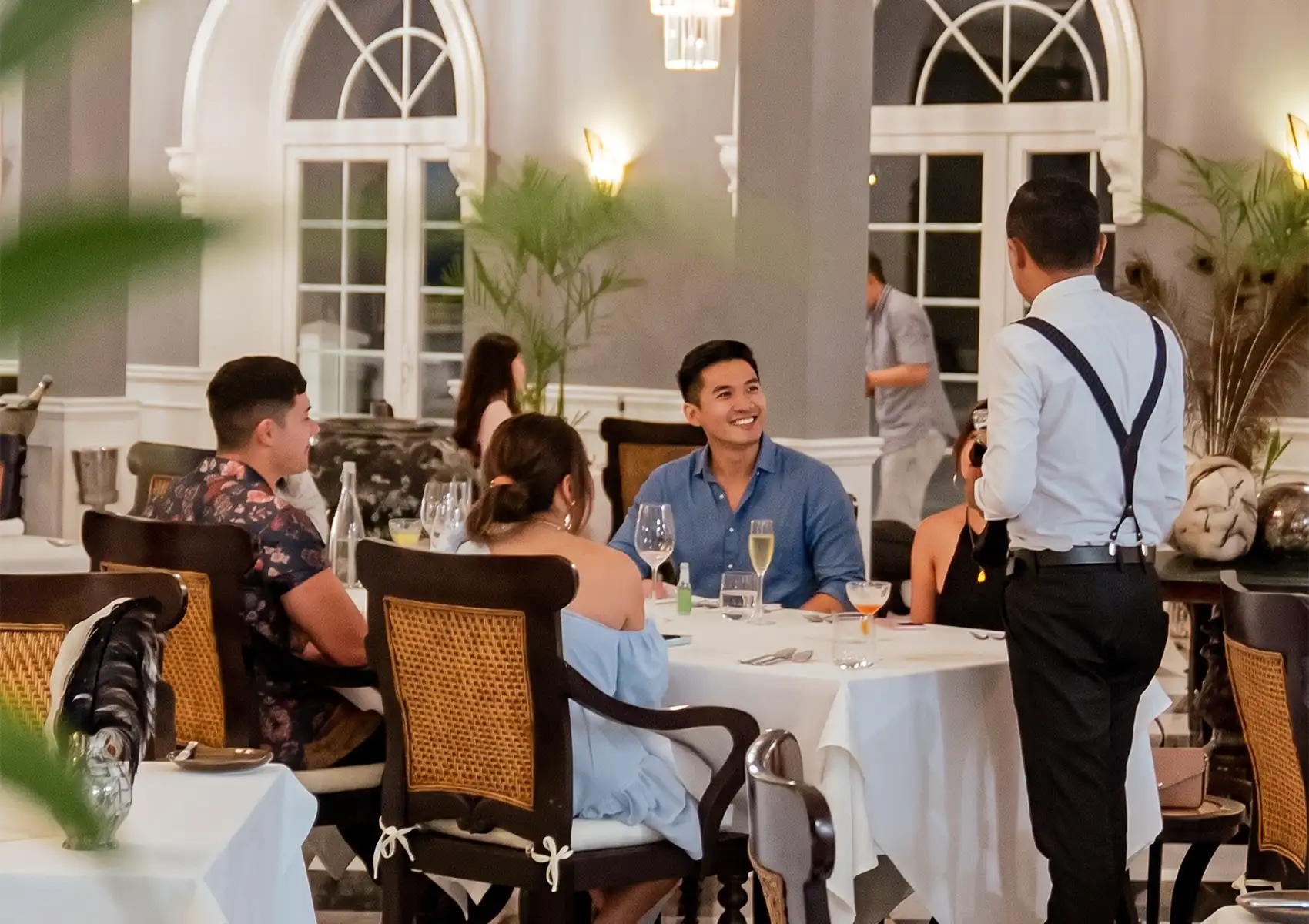 A group of elegantly dressed friends smiles and converses with a server at a beautifully set table in a spacious restaurant with large arched windows, enjoying a fine dining bali experience.
