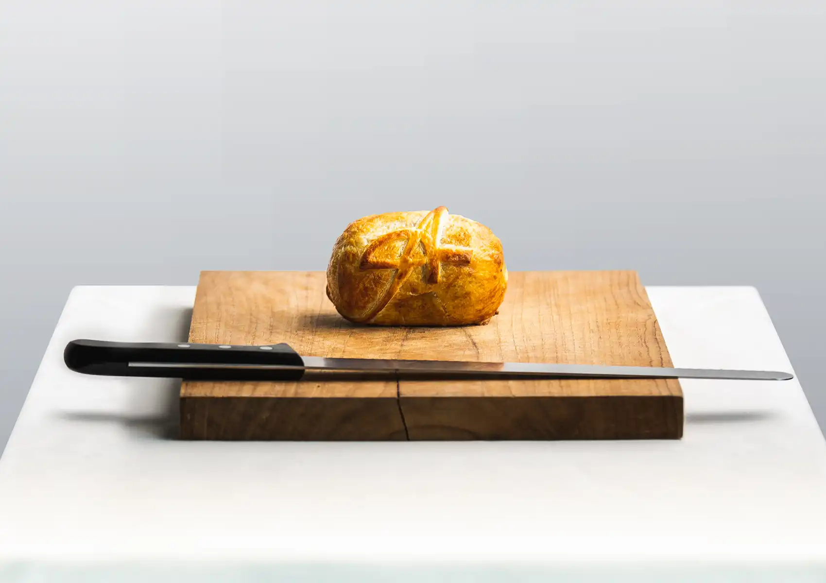 A whole, golden-brown baked Wellington pastry rests on a wooden cutting board next to a long carving knife in a minimal fine dining Bali setting.