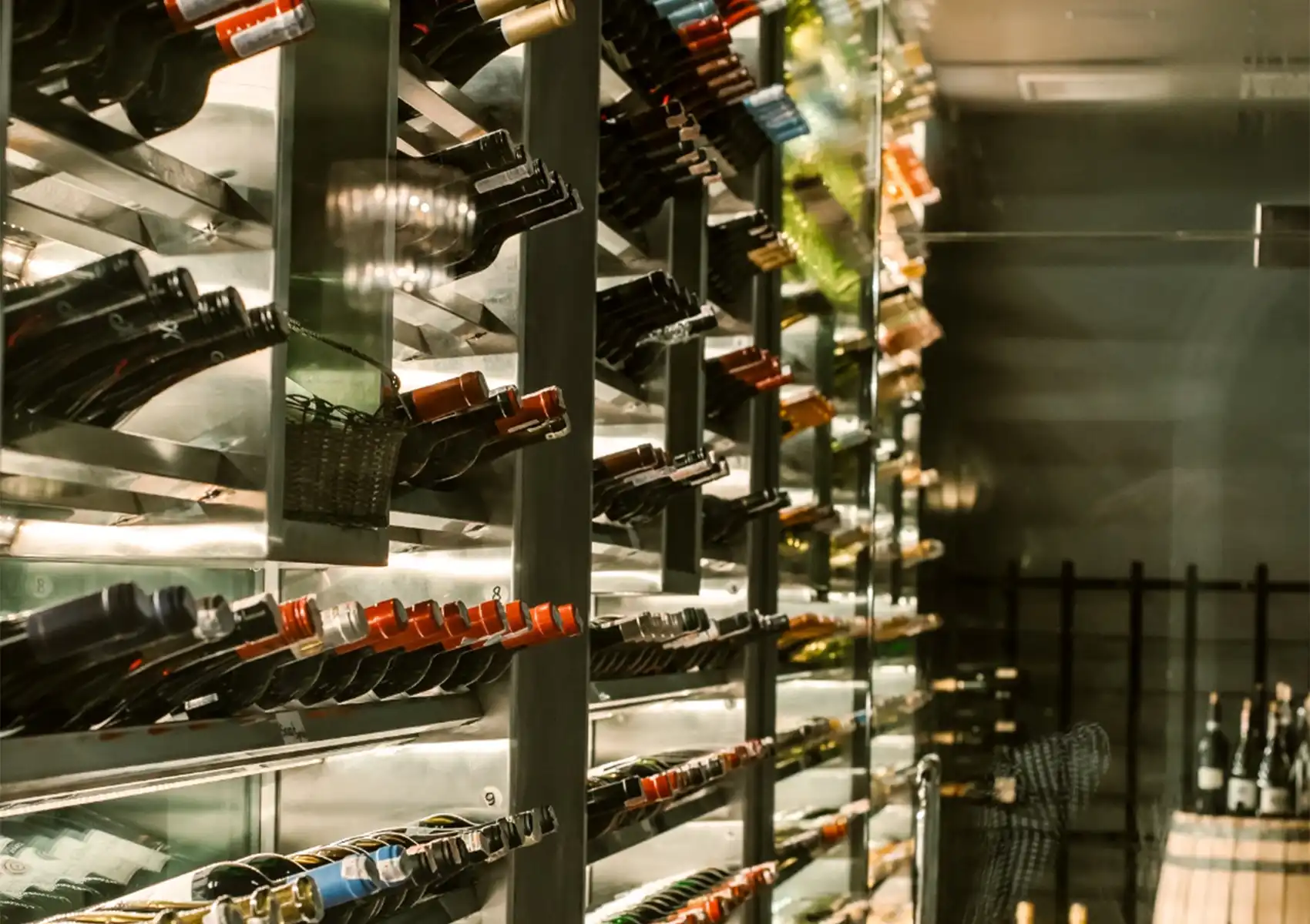 A modern, glass-enclosed wine cellar featuring neatly organized rows of wine bottles stored horizontally on metal racks, showcasing the premium beverage selection available for fine dining bali.